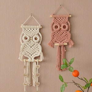 Op de afbeelding: Twee macramé wandhangers in de vorm van uilen, een in crème en een in roze, met houten latten en kwastjes. De crèmekleurige uil heeft houten kralen voor ogen en een rij houten hartjes die eronder hangen. De wandhangers worden getoond op een muur met een houten kast met geometrisch gepatroneerde deuren eronder.