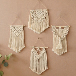 Könnte beinhalten: Vier Makramee-Wandbehänge in Cremefarbe mit Holzleisten. Die Behänge sind an einer beigefarbenen Wand mit einer weißen Lampe und einem Stuhl mit einer weißen Decke befestigt.