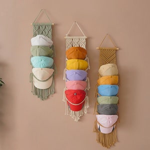 May include: Three vertical macrame wall hangings made of cord, each featuring a wooden dowel at the top and a series of loops designed to hold multiple baseball caps. The caps are displayed in various colors including white, green, blue, yellow, and red.