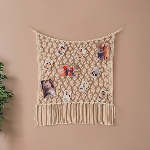 Puede incluir: Un tapiz de macramé blanco con borde de flecos, decorado con fotos. El tapiz se muestra en una pared beige en un ambiente de sala de estar con dos sillones blancos y una mesa auxiliar.