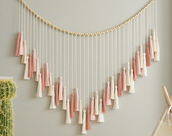 May include: A wall hanging with a string of wooden beads and pink and white tassels. The tassels are arranged in a curved shape. The wall hanging is hung on a light gray wall. The text "HEARTCRAFTED heartcraftedbyn.etsy.com" is at the bottom of the image.
