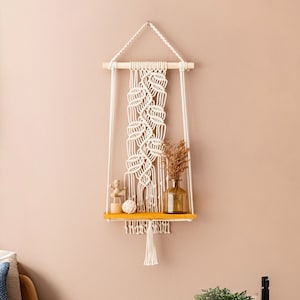 May include: A macrame wall shelf with a yellow wooden shelf and white macrame hanging from a wooden dowel. The shelf is decorated with small decorative items, including a small wooden figure, a seashell, and a glass bottle.
