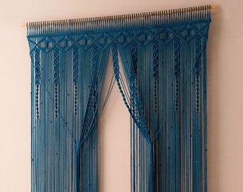 May include: A teal blue macrame curtain with a geometric pattern at the top. The curtain is made of thin cords and hangs from a wooden dowel.
