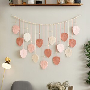 May include: A wall hanging made of woven yarn in the shape of leaves. The leaves are in shades of pink, cream, and brown. The leaves are strung together with wooden beads and twine. The wall hanging is hanging on a white wall.
