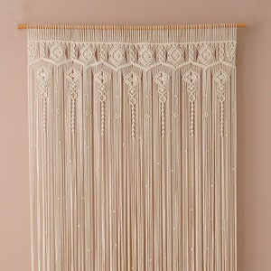 Könnte beinhalten: Ein weißer Makramee-Wandbehang mit geometrischem Muster. Der Behang ist aus Baumwollschnur gefertigt und hat oben eine Holzleiste.