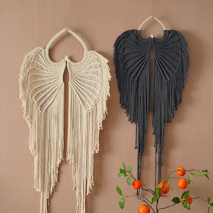 Op de afbeelding: Twee macramé wandhangers in de vorm van engelenvleugels, één in crème en één in donkergrijs, hangen aan een beige muur. Een blauwe en witte gebloemde vaas met een kleine oranjeboomtak staat op een houten tafel met een geometrisch patroon.