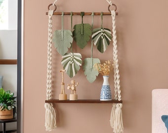 May include: A macrame wall hanging with a wooden shelf and green leaf designs. The shelf is decorated with a small vase, dried flowers, and wooden figurines.