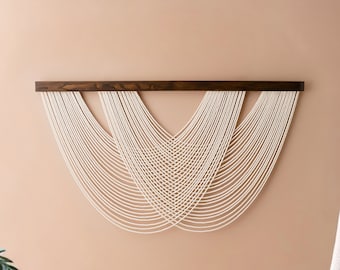 Könnte beinhalten: Ein weißer Makramee-Wandbehang mit einem Holzrahmen, der an einer hellpfirsichfarbenen Wand hängt. Der Makramee hat die Form eines Halbmondes, wobei die Schnüre in einer Wellenform nach unten fallen.
