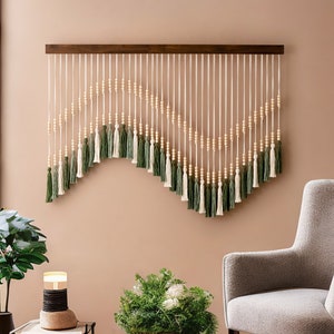 May include: A wall hanging made of white string with wooden beads and green tassels. The hanging is in the shape of a wave and is mounted on a wooden bar.
