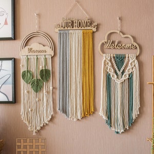 May include: Three macrame wall hangings with wooden signs. The leftmost hanging features a rainbow and cloud with the name "Vanessa" on it. The middle hanging has a sign that says "Welcome to Our Home". The rightmost hanging has a cloud sign that says "Welcome".