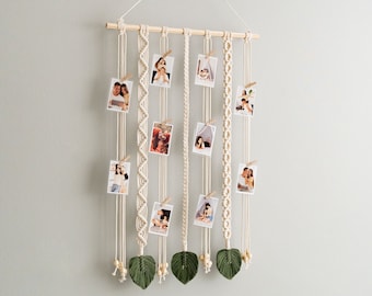 Könnte beinhalten: Ein Wandbehang aus Makramee mit einer Holzleiste oben und grünen Blättern unten. Der Wandbehang besteht aus mehreren Makramee-Strängen, an denen Fotos befestigt sind.