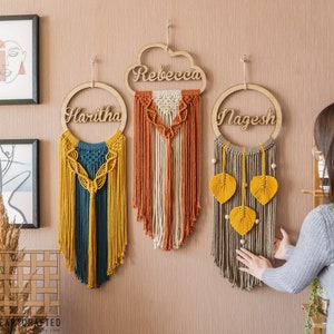 Puede incluir: Tres tapices de macramé de pared con letreros de madera en forma de nube con los nombres "Haritha", "Rebecca" y "Nagesh" escritos en ellos.