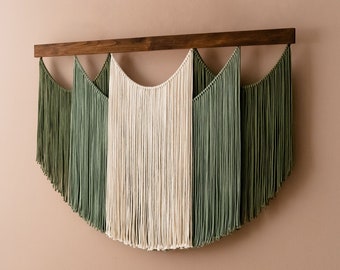 May include: A wall hanging made of yarn with a wooden dowel at the top. The yarn is in three sections, with two sections of green yarn and one section of cream yarn. The yarn is arranged in a curved shape.