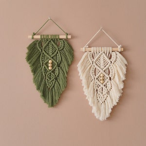 May include: Two macrame wall hangings with wooden dowels and wooden beads. The wall hanging on the left is green and the wall hanging on the right is cream.