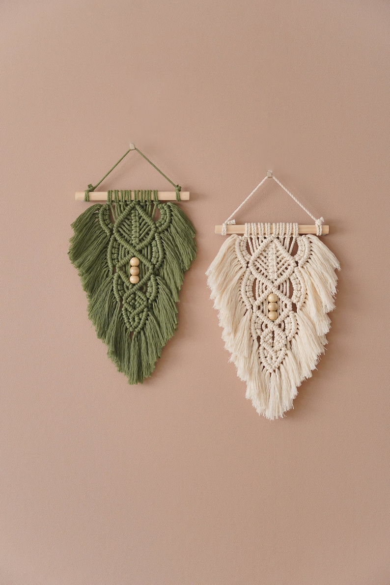 May include: Two macrame wall hangings with wooden dowels and wooden beads. The wall hanging on the left is green and the wall hanging on the right is cream.
