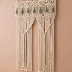 May include: A white macrame wall hanging with a geometric design and green tassels. The hanging is made of cotton cord and is attached to a wooden dowel.