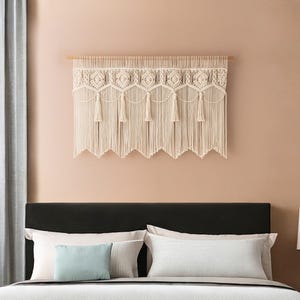 May include: Cream-colored macrame wall hanging with a geometric design and fringe. The textile is mounted on a wooden dowel and hangs above a bed with pillows. The wall is painted a light peach color.