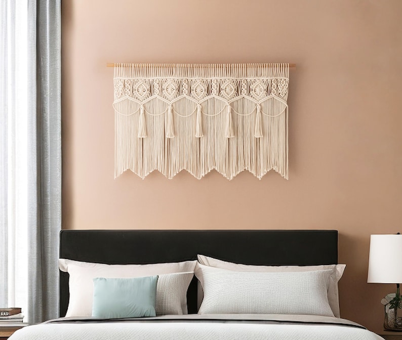 May include: Cream-colored macrame wall hanging with a geometric design and fringe. The textile is mounted on a wooden dowel and hangs above a bed with pillows. The wall is painted a light peach color.