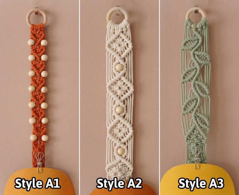 K&ouml;nnte beinhalten: Drei Makramee-Wandbeh&auml;nge mit unterschiedlichen Mustern und Farben. Der erste ist orange mit Holzperlen, der zweite ist wei&szlig; mit Holzperlen und der dritte ist gr&uuml;n. Jeder Behang hat oben einen Holzring und unten einen Metallclip. Die Beh&auml;nge sind mit "Style A1", "Style A2" und "Style A3" beschriftet.