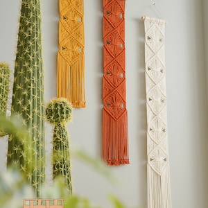 May include: Three macrame wall hangings in different colors. The leftmost hanging is yellow, the middle hanging is orange, and the rightmost hanging is white. Each hanging has a diamond pattern and fringe.