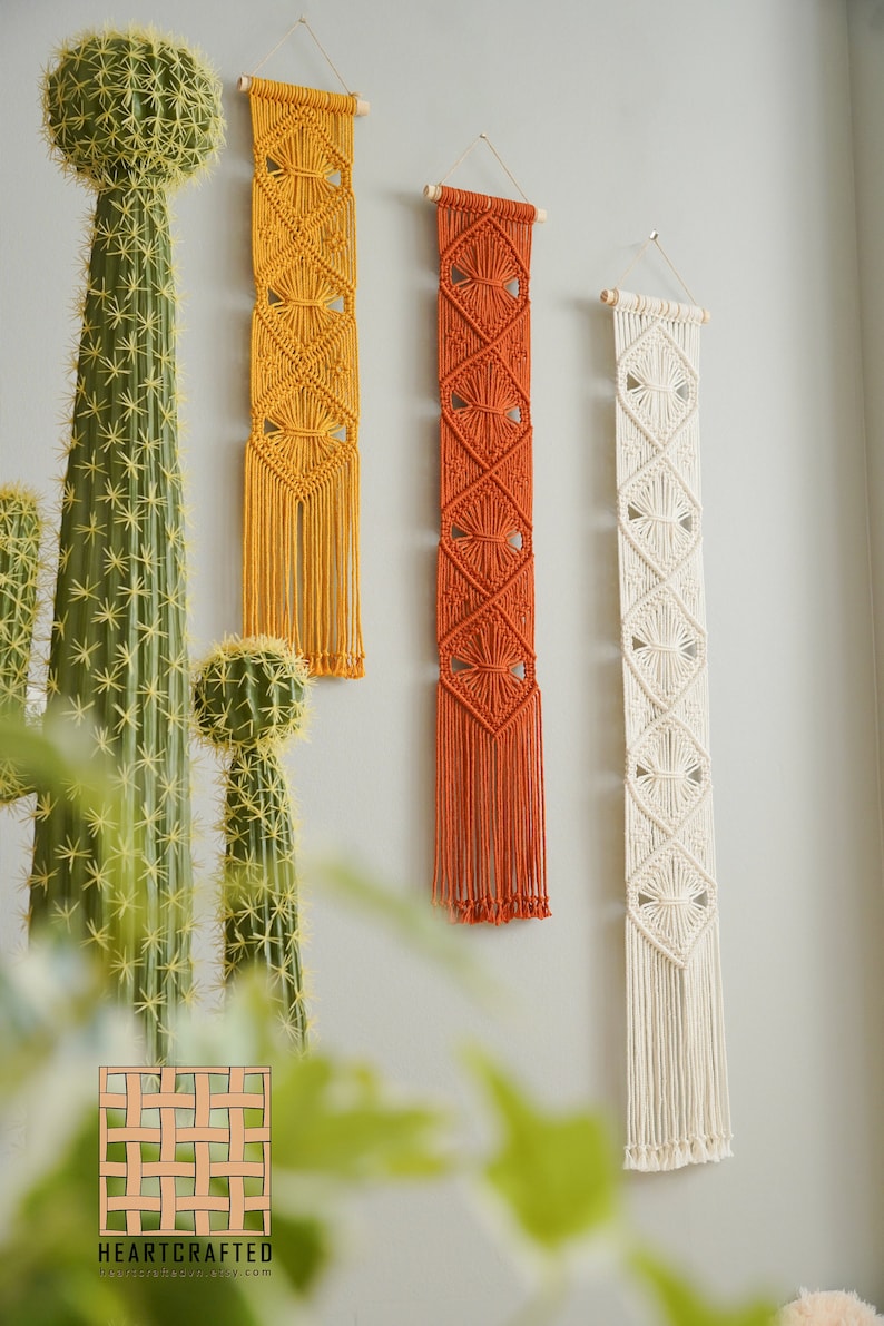May include: Three macrame wall hangings in different colors. The leftmost hanging is yellow, the middle hanging is orange, and the rightmost hanging is white. Each hanging has a diamond pattern and fringe.