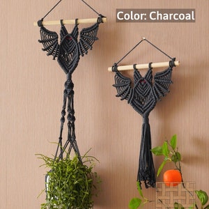 May include: Two charcoal gray macrame bat wall hangings with red eyes. The bat on the left is hanging from a plant hanger with a green plant. The bat on the right is hanging from a wooden dowel. The text "Color: Charcoal" is visible in the top right corner of the image.