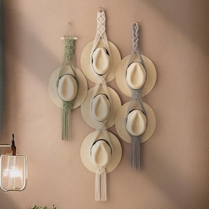 Puede incluir: Un conjunto de cinco porta sombreros de macramé con un diseño tejido. Cada porta sombrero está hecho de cuerda de algodón natural y cuenta con una repisa circular para sombreros. Los porta sombreros se cuelgan en una pared y están disponibles en tres colores: verde, gris y natural.