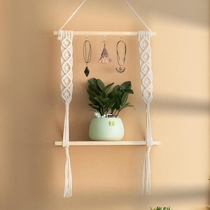 May include: A white macrame shelf with a wooden bar and two hanging strands. The shelf has a small potted plant and three hanging jewellery pieces.