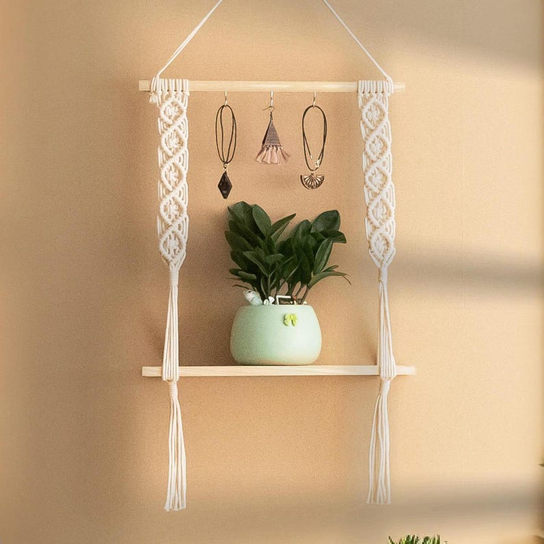 May include: A white macrame shelf with a wooden bar and two hanging strands. The shelf has a small potted plant and three hanging jewellery pieces.