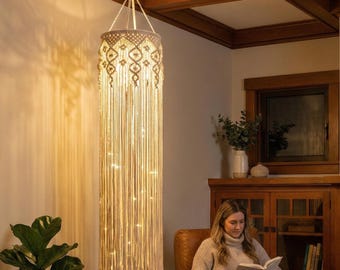 Puede incluir: Una lámpara colgante de macramé color crema con un patrón geométrico en la parte superior y largas borlas. Pequeñas luces de hadas de tonos cálidos iluminan la lámpara, creando un brillo suave. La lámpara cuelga de un gancho del techo.