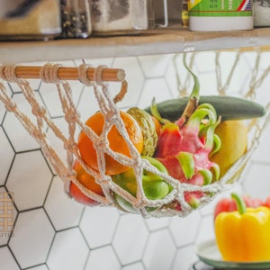 May include: A white rope hammock hanging from a wooden shelf, filled with a variety of fruits, including a dragon fruit, a green cucumber, and a yellow pear.  The hammock is hanging over a white hexagonal tile wall.