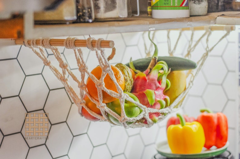May include: A white rope hammock hanging from a wooden shelf, filled with a variety of fruits, including a dragon fruit, a green cucumber, and a yellow pear.  The hammock is hanging over a white hexagonal tile wall.