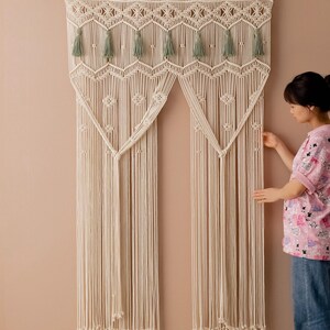 May include: A white macrame wall hanging with a geometric design and green tassels. The hanging is made of cotton cord and is approximately 3 feet wide and 4 feet tall.