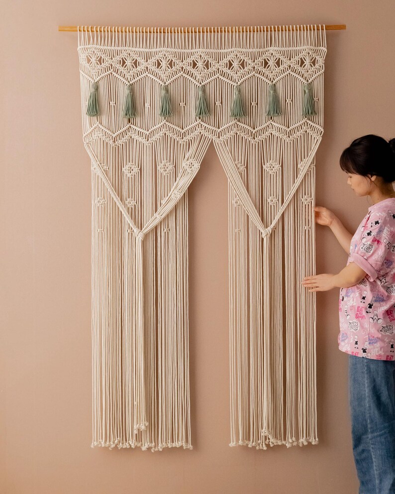 May include: A white macrame wall hanging with a geometric design and green tassels. The hanging is made of cotton cord and is approximately 3 feet wide and 4 feet tall.