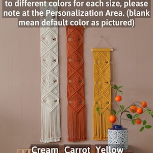 May include: Three macrame wall hangings in cream, orange, and yellow. The hangings are made with a diamond pattern and have fringe at the bottom. The text "Cream", "Carrot", and "Yellow" is written below the hangings.