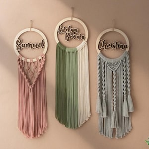 Könnte beinhalten: Drei Makramee-Wandbehänge mit Holzreifen und personalisierten Namen. Die Namen sind Evelyn Brown, Samuel und Christina. Die Behänge sind in Grün-, Rosa- und Grautönen.