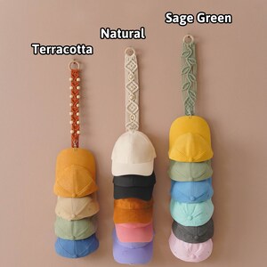K&ouml;nnte beinhalten: Eine Wandanzeige von Baseballkappen, die nach Farbe sortiert sind. Die Kappen h&auml;ngen an Makramee-Wandhaltern in Terrakotta, Natur und Salbeigr&uuml;n. Die Kappen sind in Stapeln von 7 angeordnet, wobei jeder Stapel eine andere Farbe aufweist: Gelb, Beige, Gr&uuml;n, Blau, Grau, Rosa und Lila.