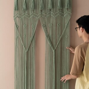 May include: A sage green macrame wall hanging with a geometric pattern and tassels. The hanging is made of cotton cord and is approximately 70 inches tall.