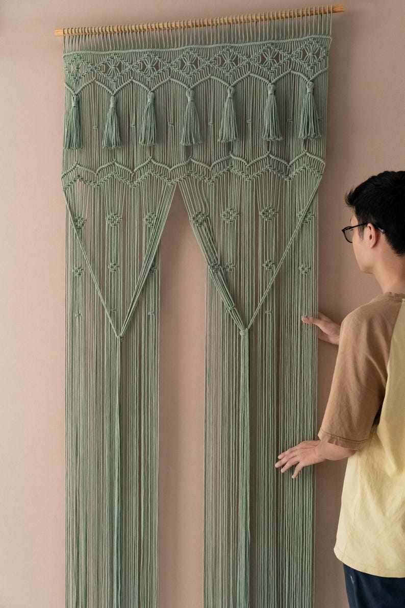 May include: A sage green macrame wall hanging with a geometric pattern and tassels. The hanging is made of cotton cord and is approximately 70 inches tall.