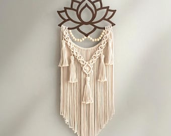 Puede incluir: Un tapiz de pared de macramé color crema con un diseño de flor de loto marrón en la parte superior. El macramé incluye borlas y cuentas de madera, creando una estética bohemia. La pieza está suspendida por una fina cuerda.