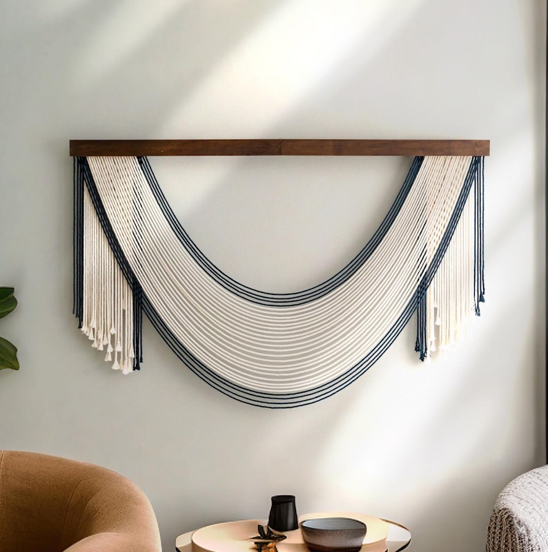 May include: A white and navy blue macrame wall hanging with a wooden dowel at the top. The macrame is in the shape of a crescent moon.
