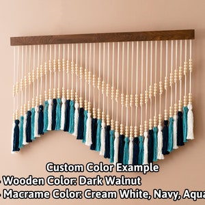 May include: A wooden wall hanging with a dark walnut finish features a macrame design with cream white, navy, and aqua tassels. The macrame is strung with wooden beads and hangs from a wooden dowel.