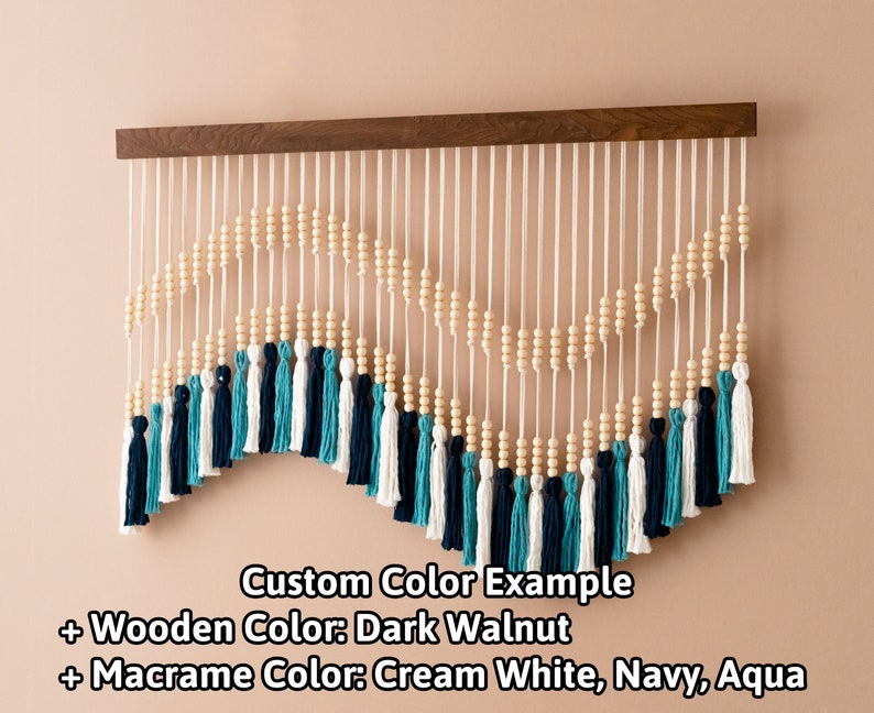 May include: A wooden wall hanging with a dark walnut finish features a macrame design with cream white, navy, and aqua tassels. The macrame is strung with wooden beads and hangs from a wooden dowel.