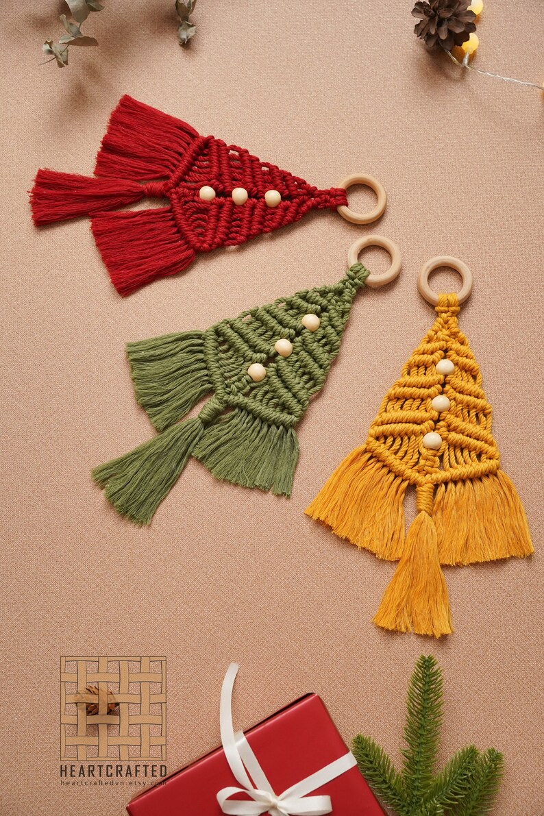 Puede incluir: Tres adornos de &aacute;rbol de Navidad de macram&eacute; hechos a mano en rojo, verde y amarillo con cuentas y anillos de madera. Los adornos est&aacute;n colgados sobre un fondo marr&oacute;n.
