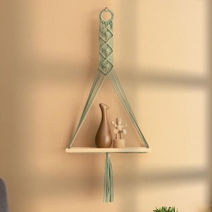 May include: A macrame shelf with a light green cord and a wooden shelf. The shelf is hanging from the ceiling and has a small wooden figurine and a brown ceramic vase on it.