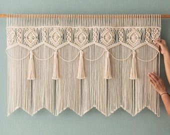 May include: Cream-colored macrame wall hanging with a geometric design and tassels. The macrame is suspended from a wooden rod, creating a decorative and textured wall accent. The design features intricate knots and a scalloped bottom edge.