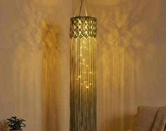 Puede incluir: Una lámpara colgante de macramé con forma cilíndrica. La sección superior presenta un diseño estampado, mientras que la parte inferior está adornada con largas y fluidas flecos. Pequeñas luces están intercaladas, creando un brillo cálido. La lámpara está suspendida de un gancho de techo.