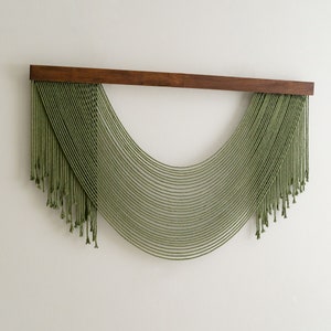 May include: A green macrame wall hanging with a wooden dowel at the top. The macrame is in the shape of a half circle with long strands of yarn hanging down.