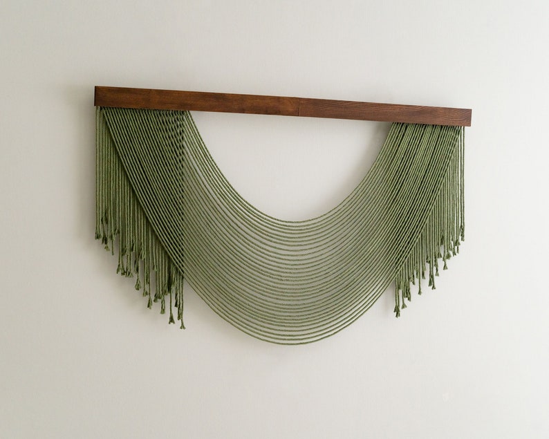 May include: A green macrame wall hanging with a wooden dowel at the top. The macrame is in the shape of a half circle with long strands of yarn hanging down.
