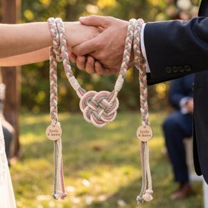 May include: A handfasting cord made of braided sage green, dusty pink, and cream rope featuring a central heart-shaped knot. Two round wooden tags are attached to the ends, each engraved with the text Emily & James in a clean font.